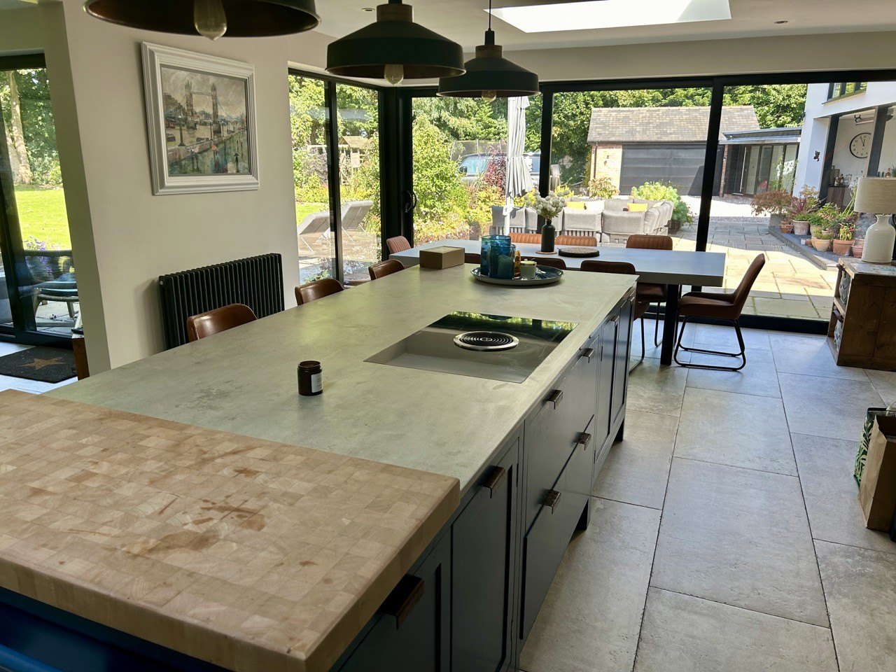 A kitchen with a long island and stone worktop under pendant lights, open to the garden